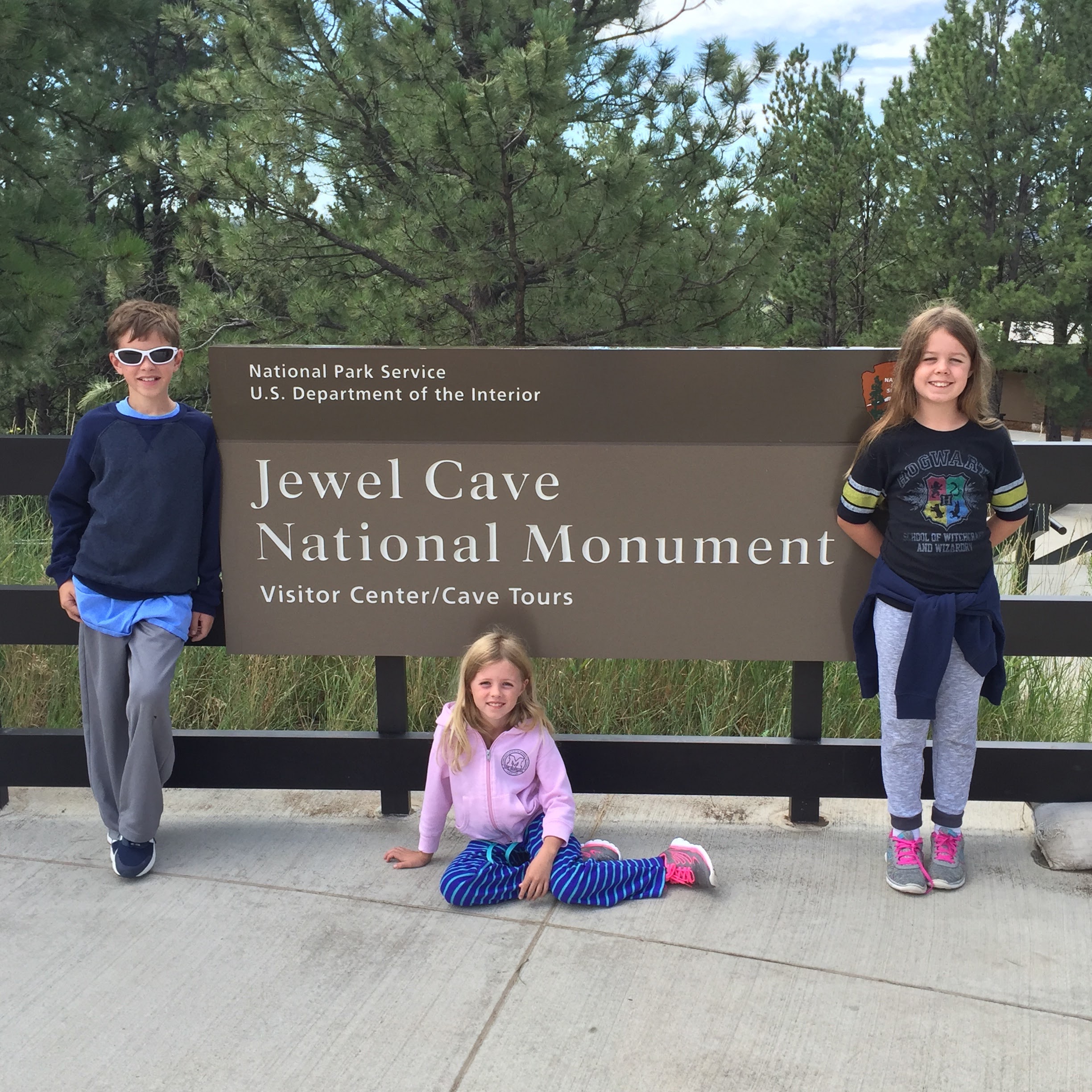 A boy and two girls standing seperately in front of sign for Jewel Cave National Momument